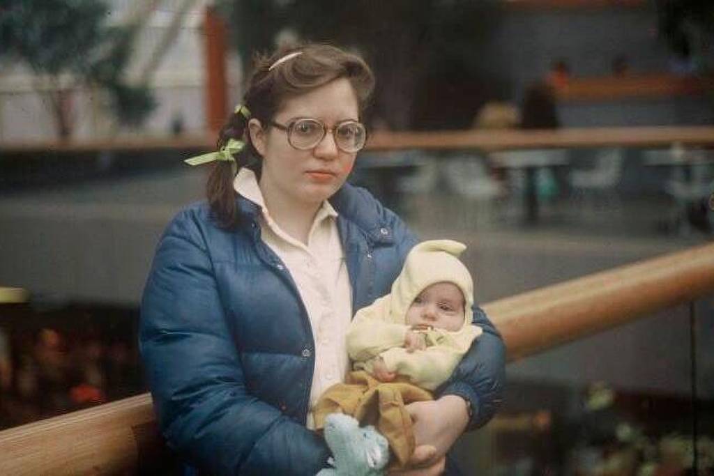 Mulher de óculos e casaco azul segura bebê vestido com roupa amarela em ambiente interno com corrimão de madeira ao fundo.