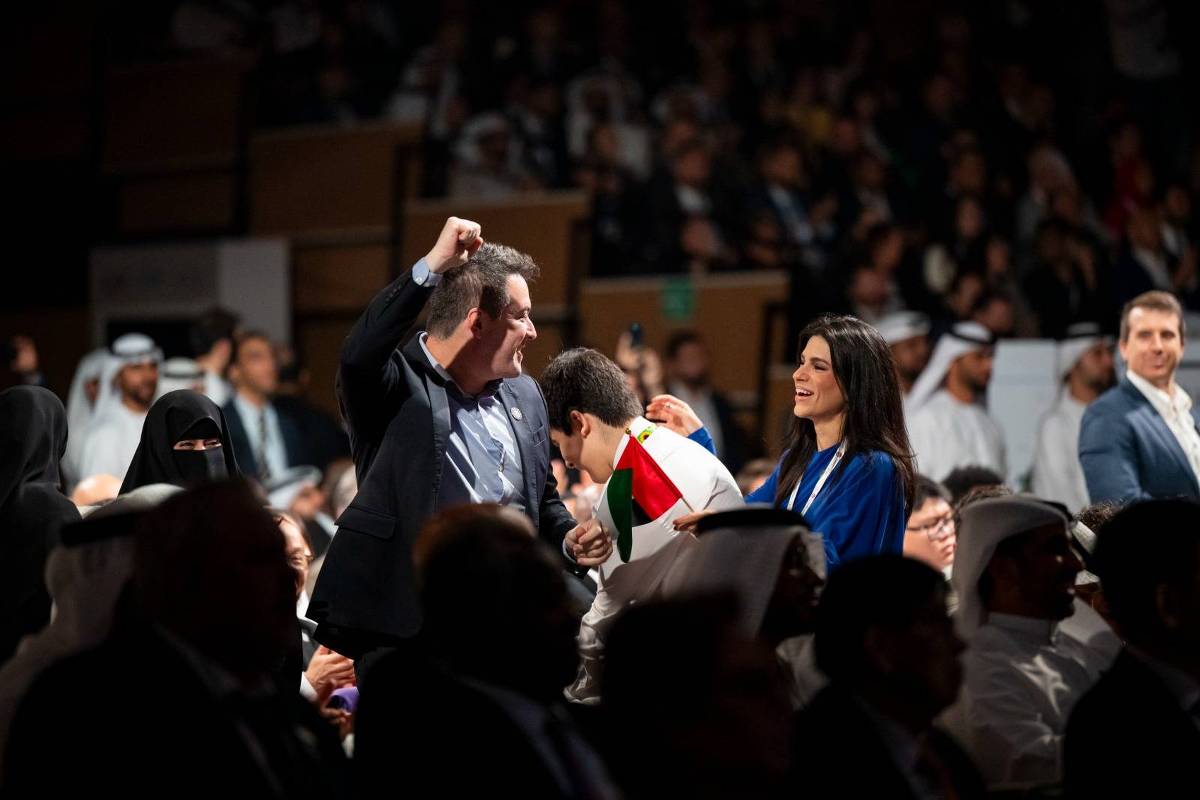 Homem levanta o punho em comemoração enquanto outro beija sua mão, com mulher sorridente ao lado. Público ao fundo inclui pessoas com trajes tradicionais árabes e ocidentais em ambiente fechado.