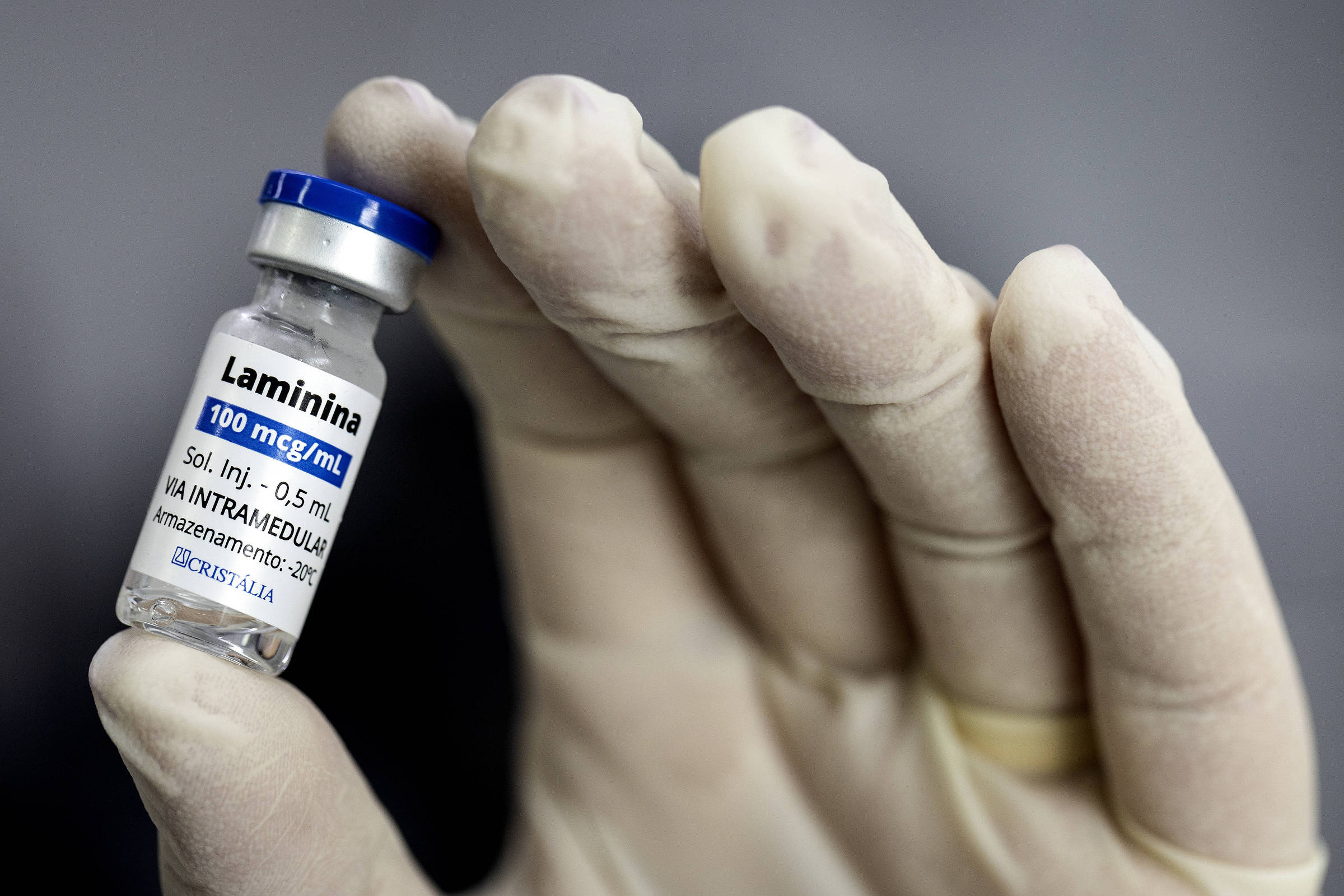Mão com luva branca segura frasco pequeno de medicamento Lamínina 100 mcg/mL, solução injetável de 0,5 mL para administração intramedular.