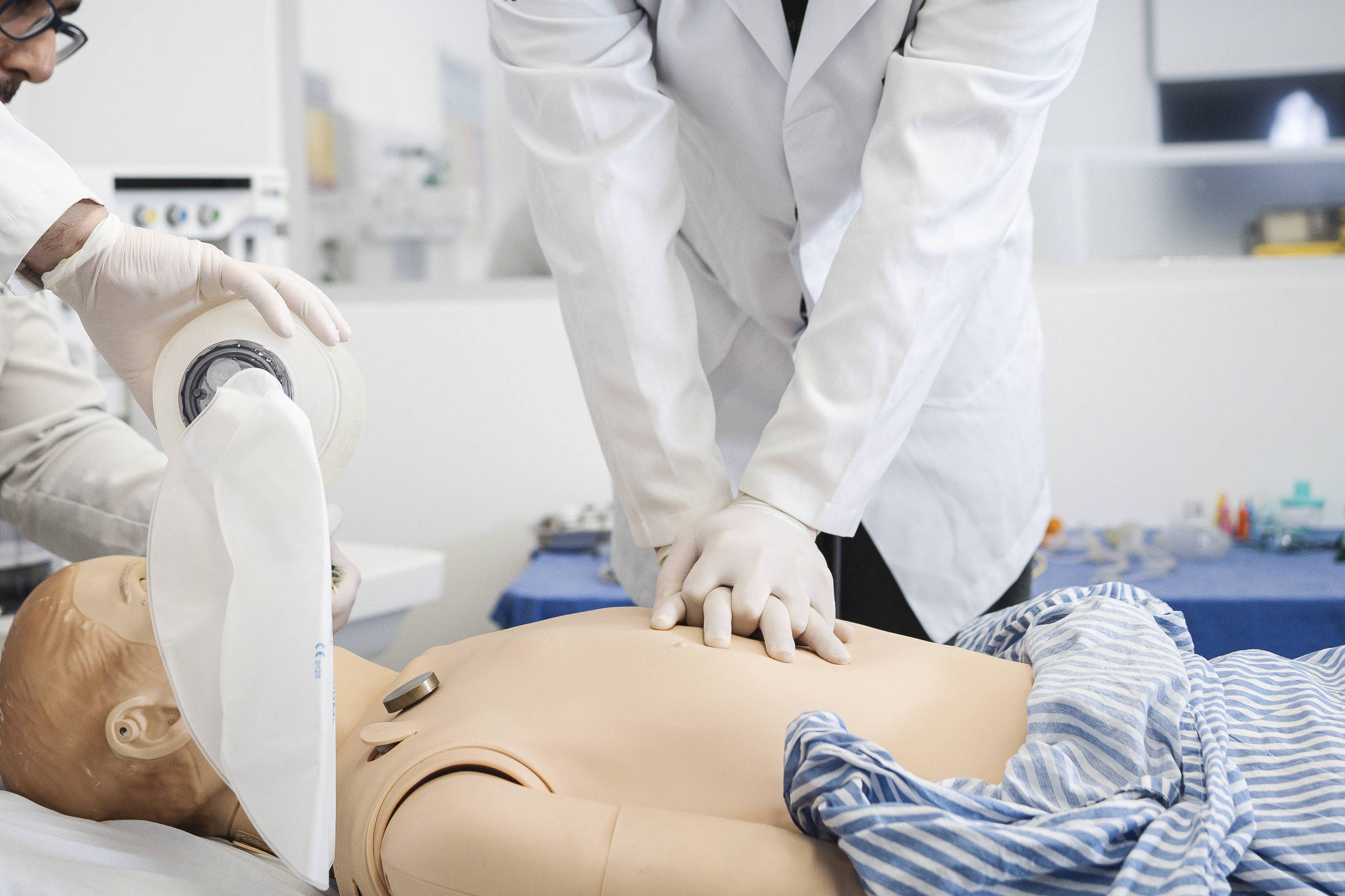 Dois profissionais de saúde em jalecos brancos realizam treinamento de ressuscitação cardiopulmonar em manequim. Um aplica compressões torácicas enquanto o outro utiliza bolsa de ventilação manual.