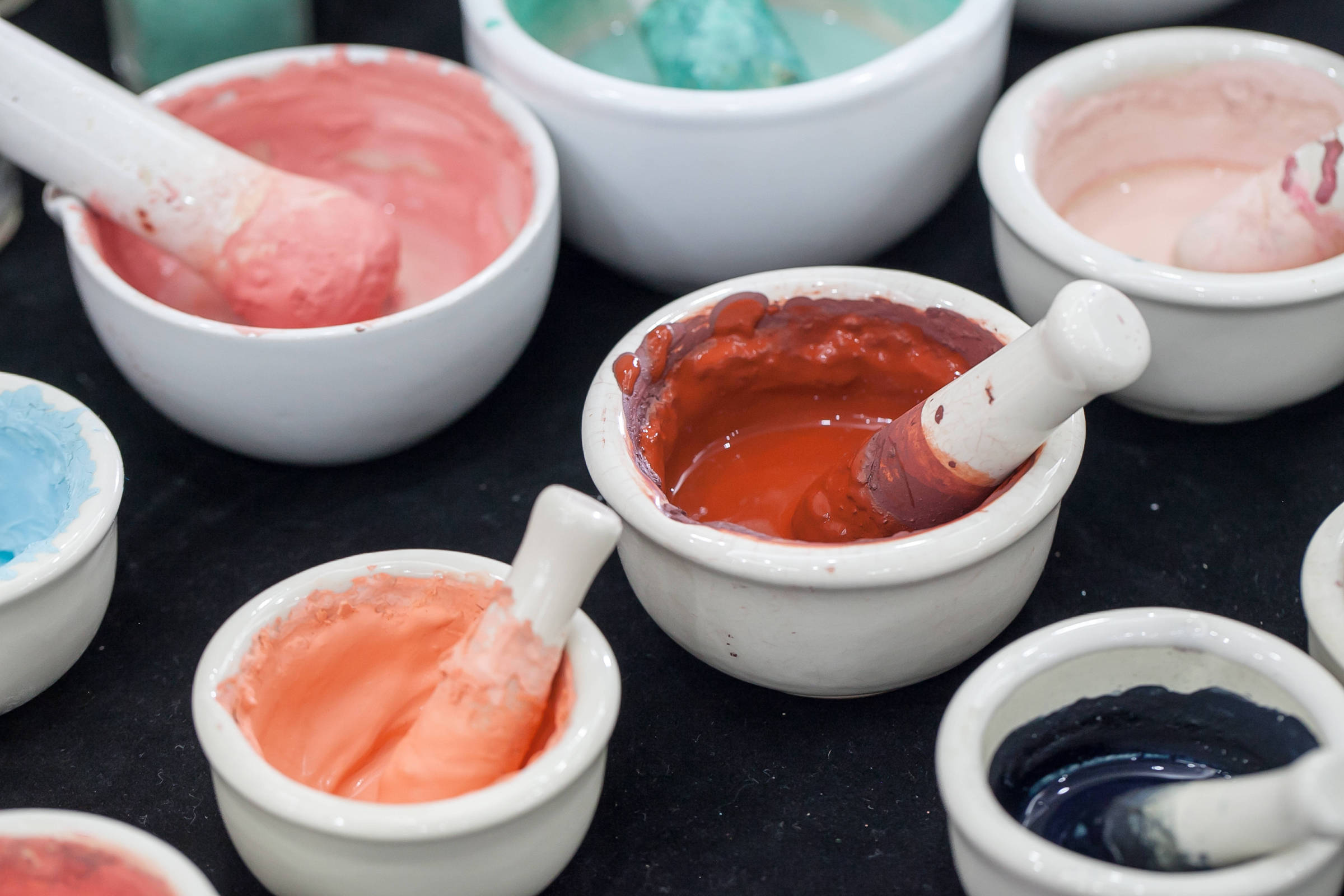 Várias tigelas brancas contendo tintas em tons de vermelho, rosa, azul e preto, cada uma com um pilão para mistura. As tigelas estão dispostas sobre uma superfície escura.