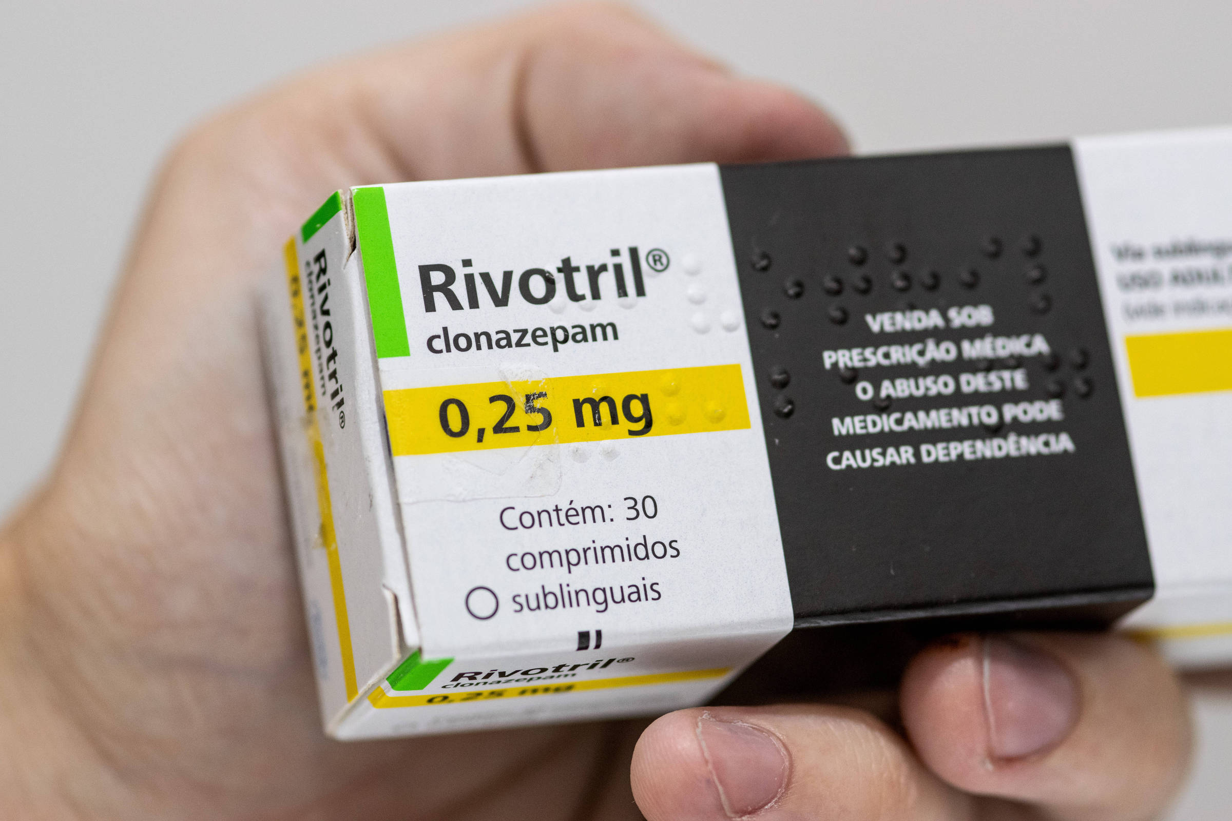 Uma mão segurando uma embalagem de Rivotril, que é um medicamento. A embalagem é branca com detalhes em verde e amarelo. Na parte frontal, está escrito